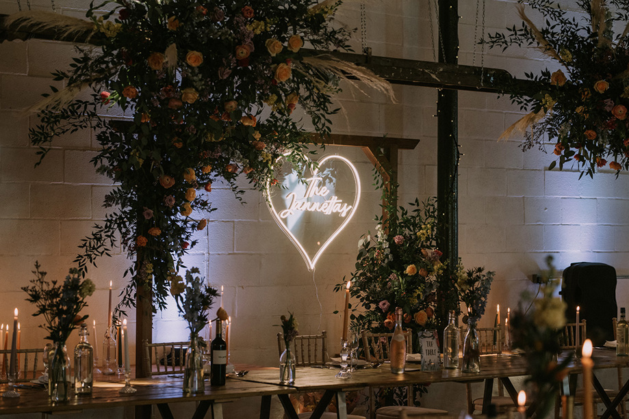 View of wooden arch decorated with wildflowers with couples surname inside love heart light in background